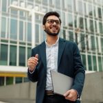 Smiling businessman in suit with laptop near modern office building is looking at side