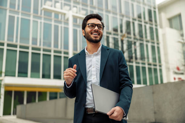 Smiling businessman in suit with laptop near modern office building is looking at side